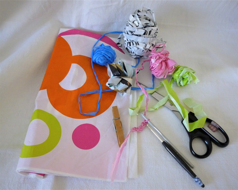 Sac plastique avec des cercles de couleurs, cinq pelotes de fils de plastique, une paire de ciseau, deux crochets et une pince à linge sur un fond blanc